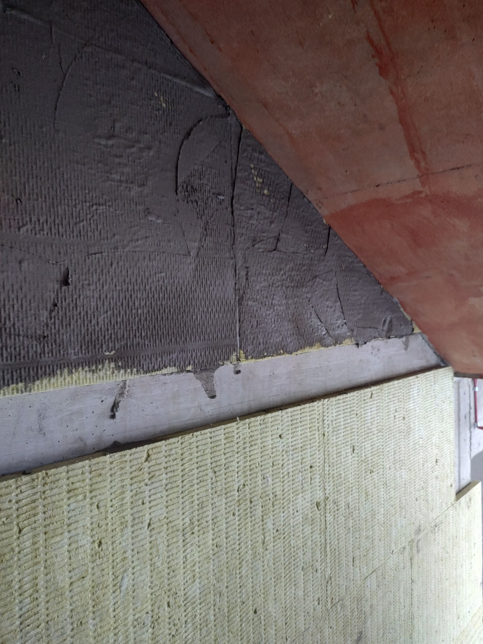 Ocieplanie ściany wełną mineralną z widoczną warstwą szpachli, w trakcie prac izolacyjnych w korytarzu bloku mieszkalnego. Widoczna struktura materiałów.