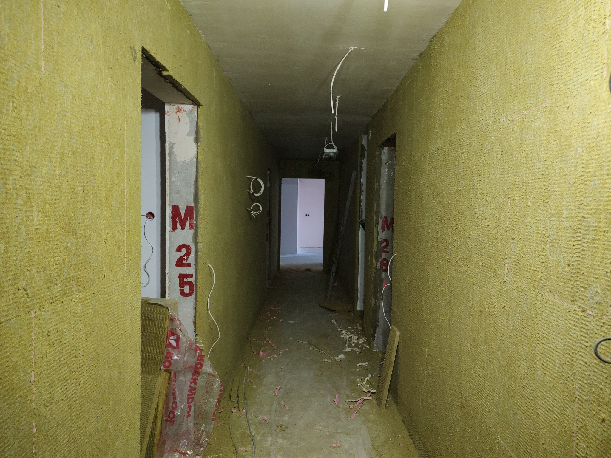 Korytarz w bloku mieszkalnym w trakcie izolacji ścian wełną mineralną. Widoczne fragmenty instalacji elektrycznej i oznaczenia 'M 25' na ścianie.