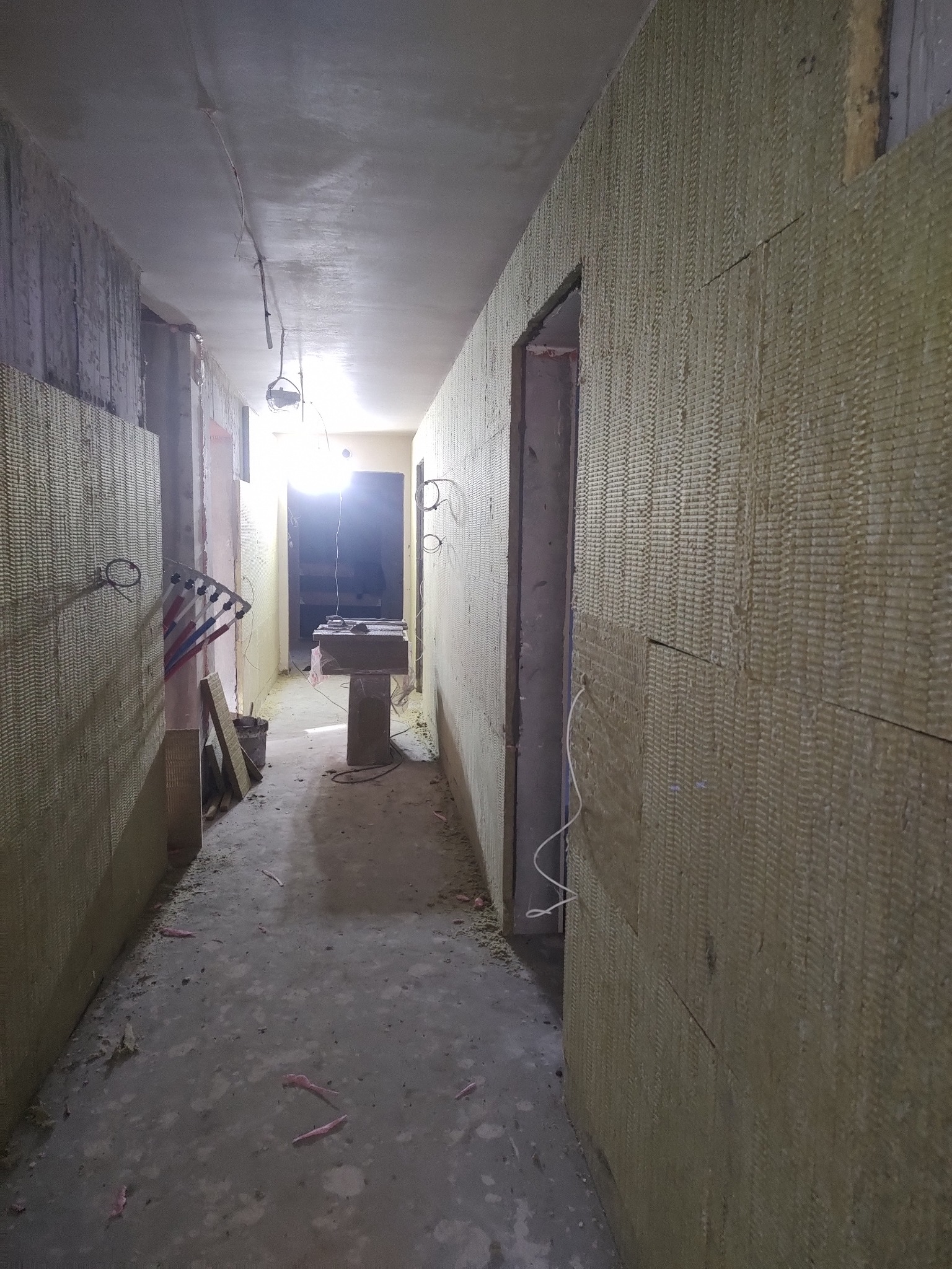 Korytarz w trakcie ocieplania wełną mineralną. Widoczne ściany pokryte materiałem izolacyjnym, surowy beton na suficie i podłodze. Etap prac wykończeniowych.