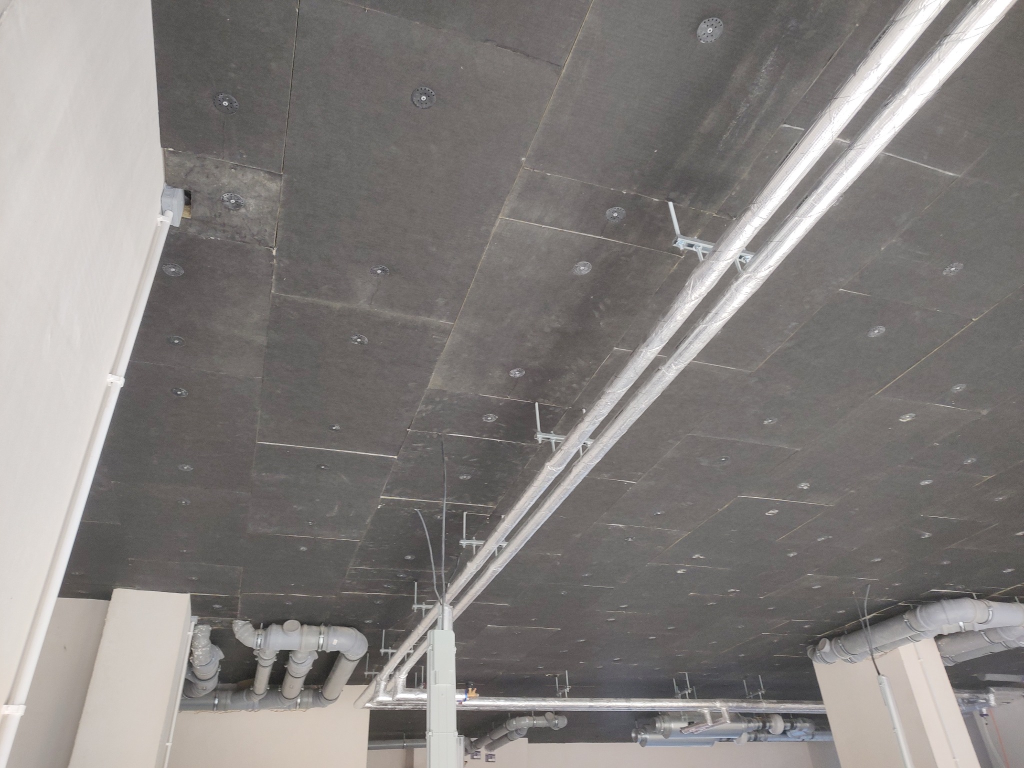 Ocieplony strop wełną mineralną z widocznymi mocowaniami i instalacją wentylacyjną w lokalu usługowym. Czarny welon na ociepleniu.