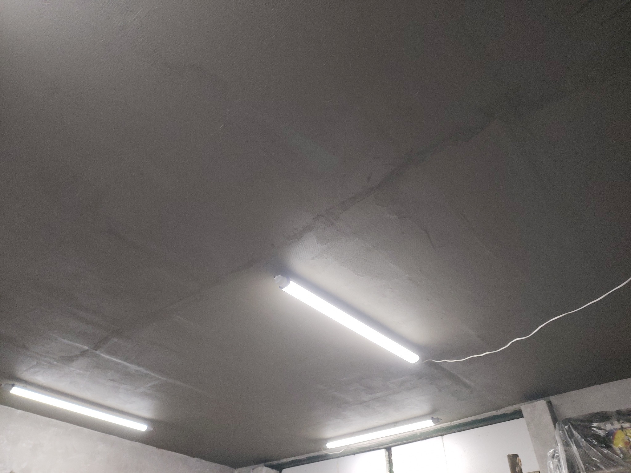 Szary sufit po szpachlowaniu i wygładzeniu z widocznymi liniami łączeń płyt i dwoma lampami jarzeniowymi, wykonany w garażu.