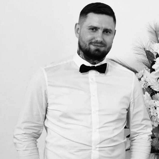 Czarno-biały portret mężczyzny w białej koszuli i muszce na tle jasnej ściany z dekoracją kwiatową, widoczny tatuaż na szyi.