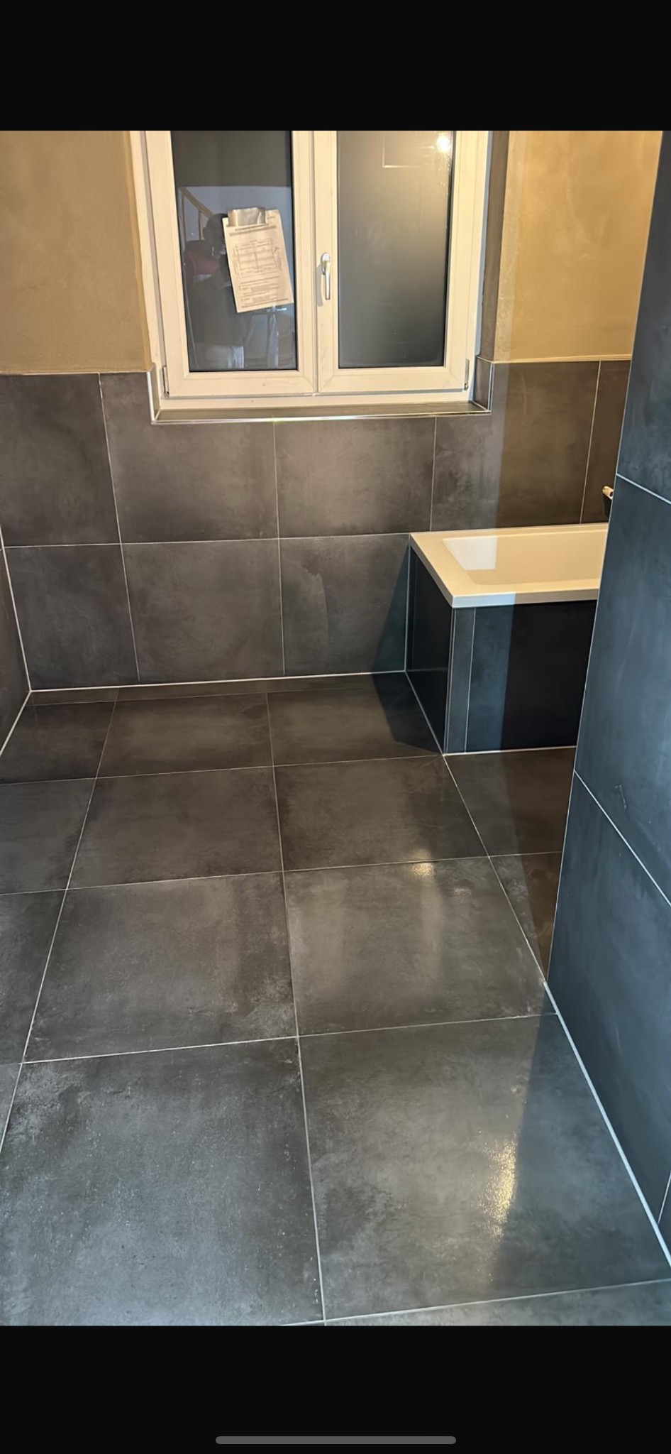 Wykończona łazienka z ciemnymi, kwadratowymi płytkami na podłodze i ścianach, jasną fugą i widoczną wanną. Na oknie wisi kartka z dokumentami.