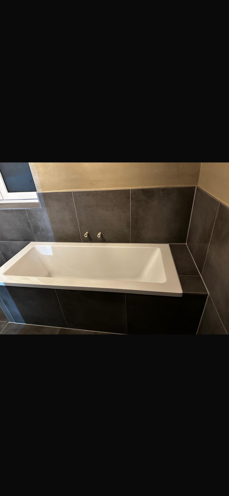 Nowoczesna, prostokątna wanna obudowana ciemnymi płytkami w łazience. Widoczne dwa wystające zawory na ścianie i fragment okna. Minimalistyczny design.