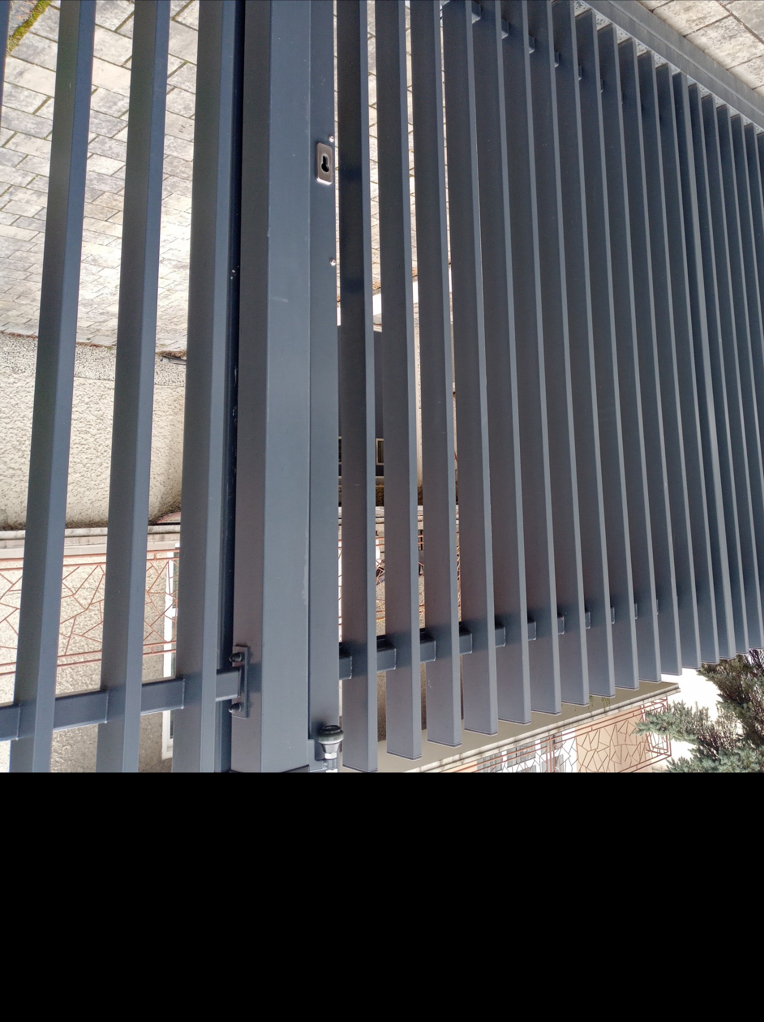 Nowoczesna, grafitowa brama z pionowych, prostokątnych profili, widoczny zamek i fragment posesji w tle. Minimalistyczny design i solidne wykonanie.