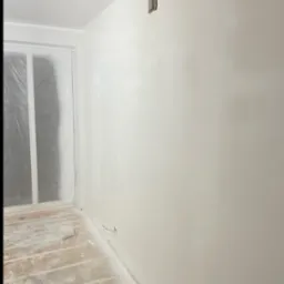 Wnętrze po gładziowaniu ścian na biało. Widoczne zabezpieczone folią okno, gniazdko elektryczne i częściowo zakryta podłoga. Świeżo wykończone, jasne pomieszczenie.