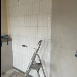 Ściana z białymi, wąskimi płytkami ułożonymi pionowo w łazience, widoczne poziomice i drabina, w trakcie prac wykończeniowych.