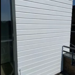 Biała, drewniana elewacja budynku z oknem i fragmentem balustrady. Widoczne poziome deski z fakturą drewna i świeże, jednolite malowanie.