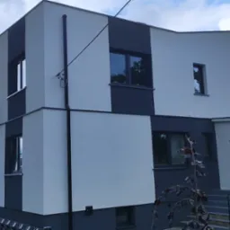 Dwukondygnacyjny dom z balkonem, pomalowany w odcieniach szarości, z widoczną elewacją i fragmentem ogrodzenia. Ujęcie z dołu, w tle zieleń drzew.