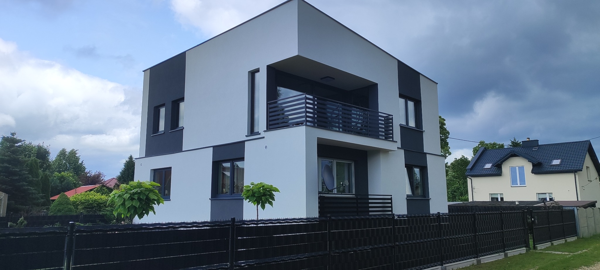 Nowoczesny dom z biało-grafitową elewacją, widok z dołu, czarne balustrady balkonów, ciemne ogrodzenie, zieleń w tle, pochmurne niebo.