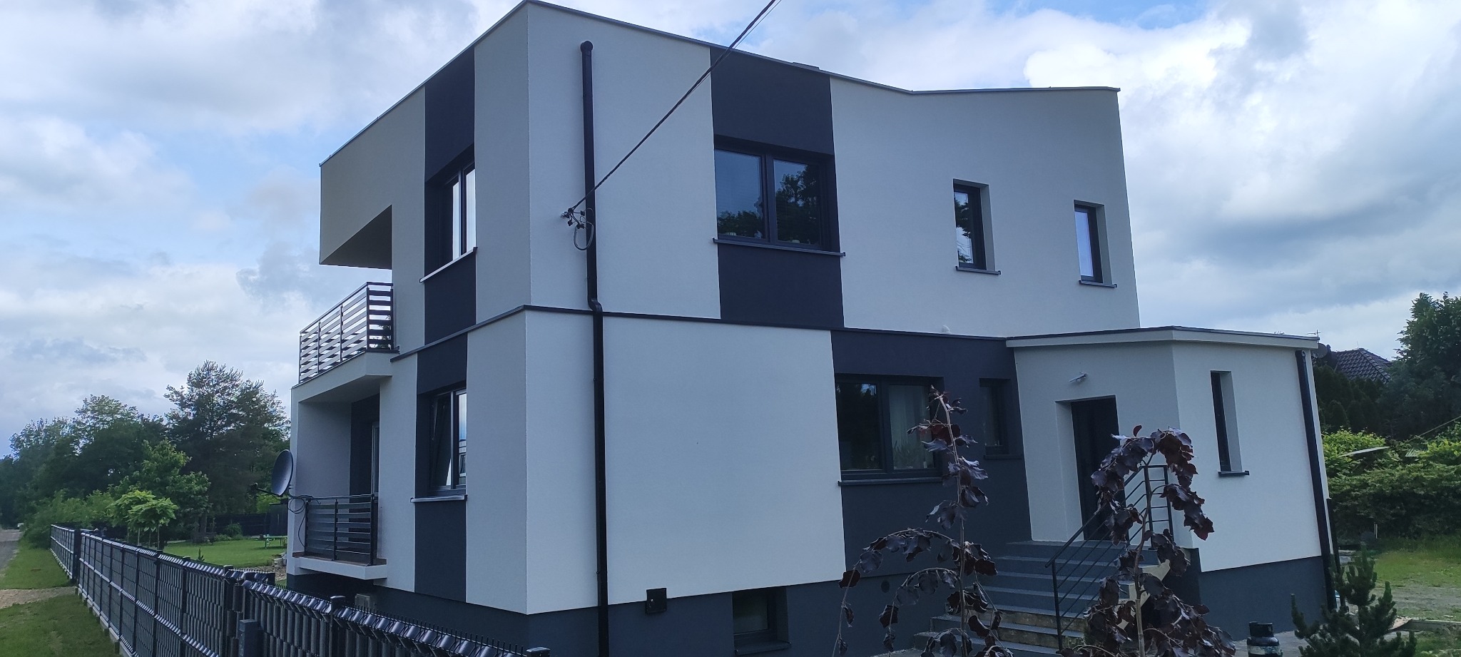Dwukondygnacyjny dom z balkonem, pomalowany w odcieniach szarości, z widoczną elewacją i fragmentem ogrodzenia. Ujęcie z dołu, w tle zieleń drzew.