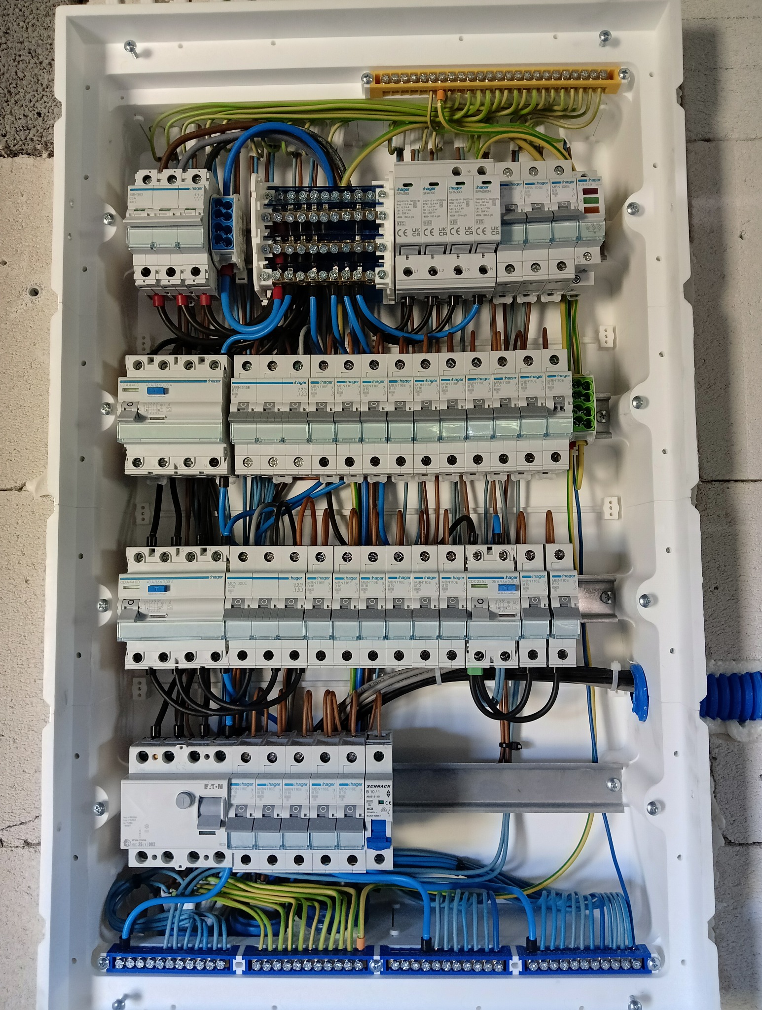 Rozdzielnica elektryczna w białej obudowie z licznymi bezpiecznikami, przewodami w kolorach niebieskim, brązowym, zielono-żółtym. Widoczne elementy montażowe.