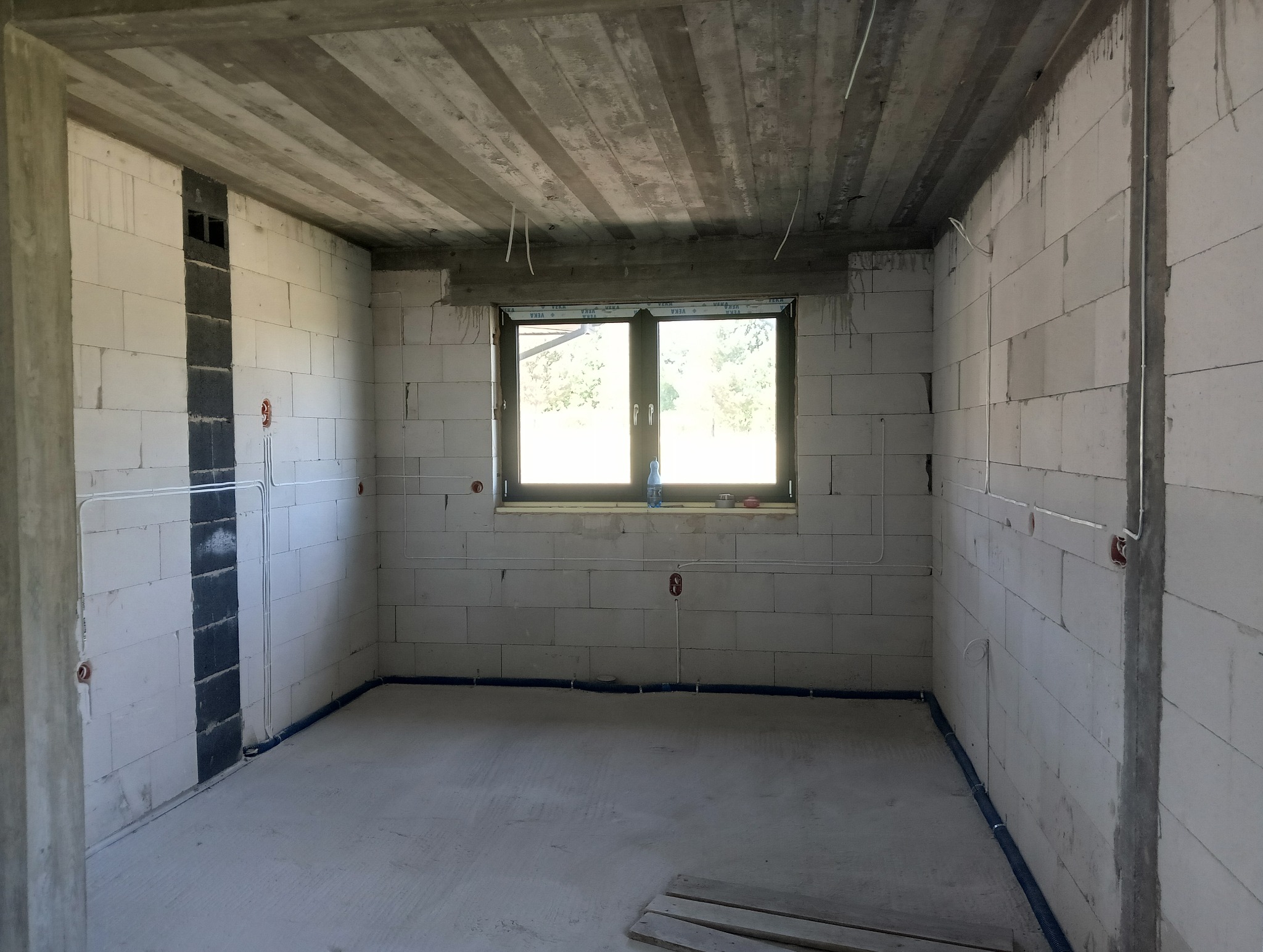 Surowy pokój z instalacją elektryczną w ścianach z betonu komórkowego. Widoczne okno, kable i rury instalacyjne. W tle drzewa.