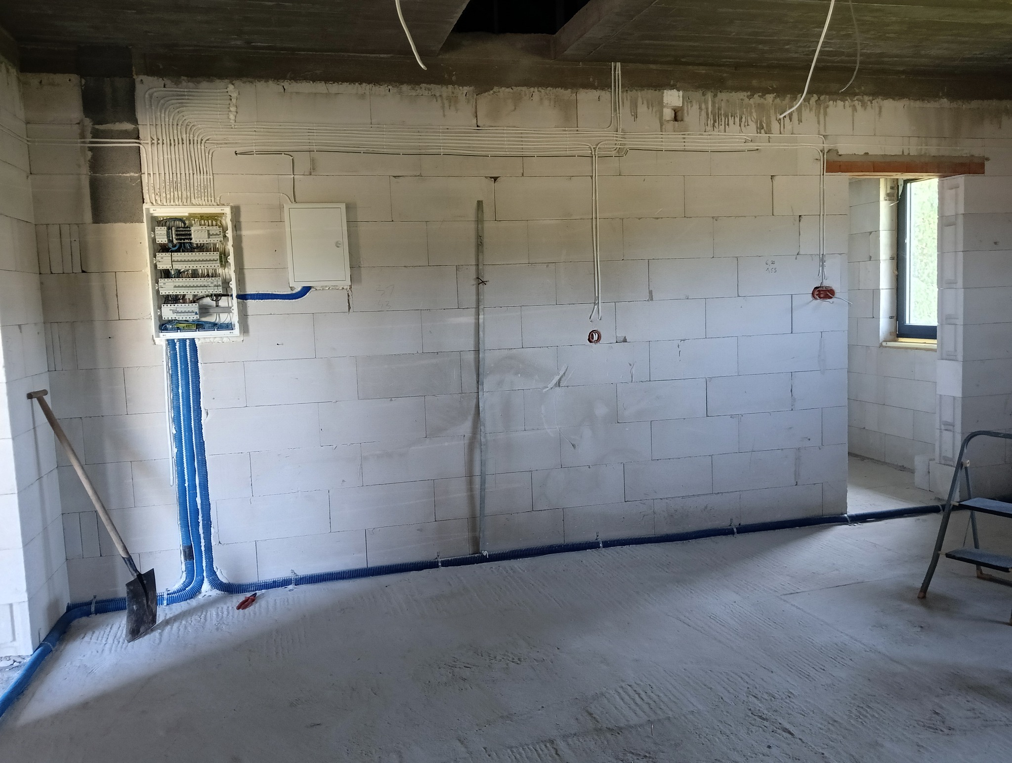 Rozdzielnia elektryczna z niebieskimi rurami osłonowymi na ścianie z betonu komórkowego w surowym wnętrzu budynku w trakcie prac instalacyjnych.
