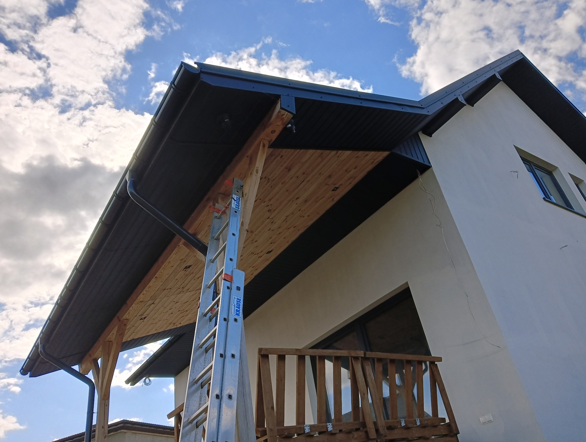 Montaż podbitki dachowej z drewnianą boazerią widoczny z dołu, z drabiną opartą o konstrukcję. Czarny system rynnowy i błękitne niebo w tle.