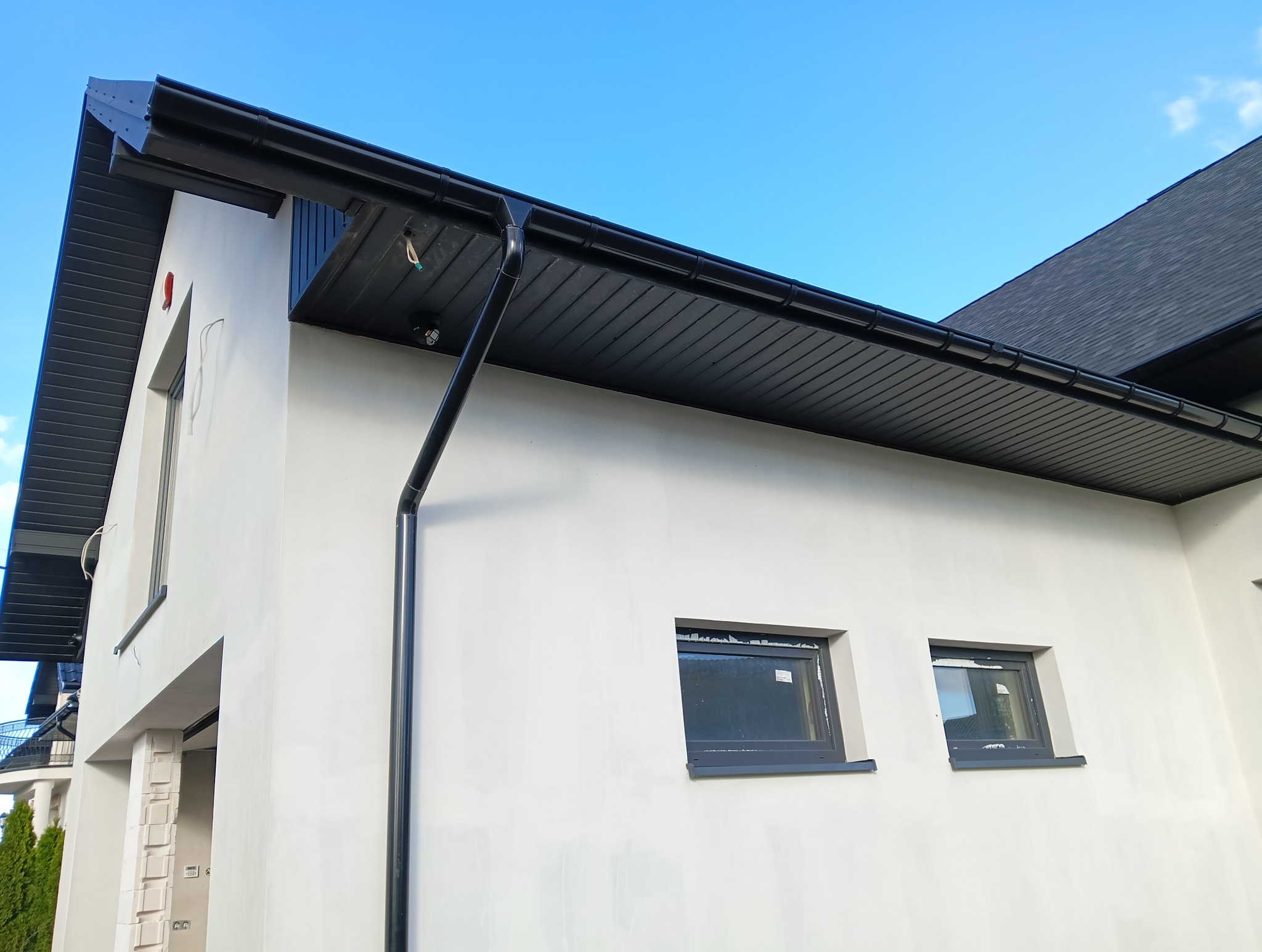 Elewacja domu jednorodzinnego z czarnym orynnowaniem i podbitką. Widoczne okna z szarą stolarką. Na elewacji zamontowany czujnik.