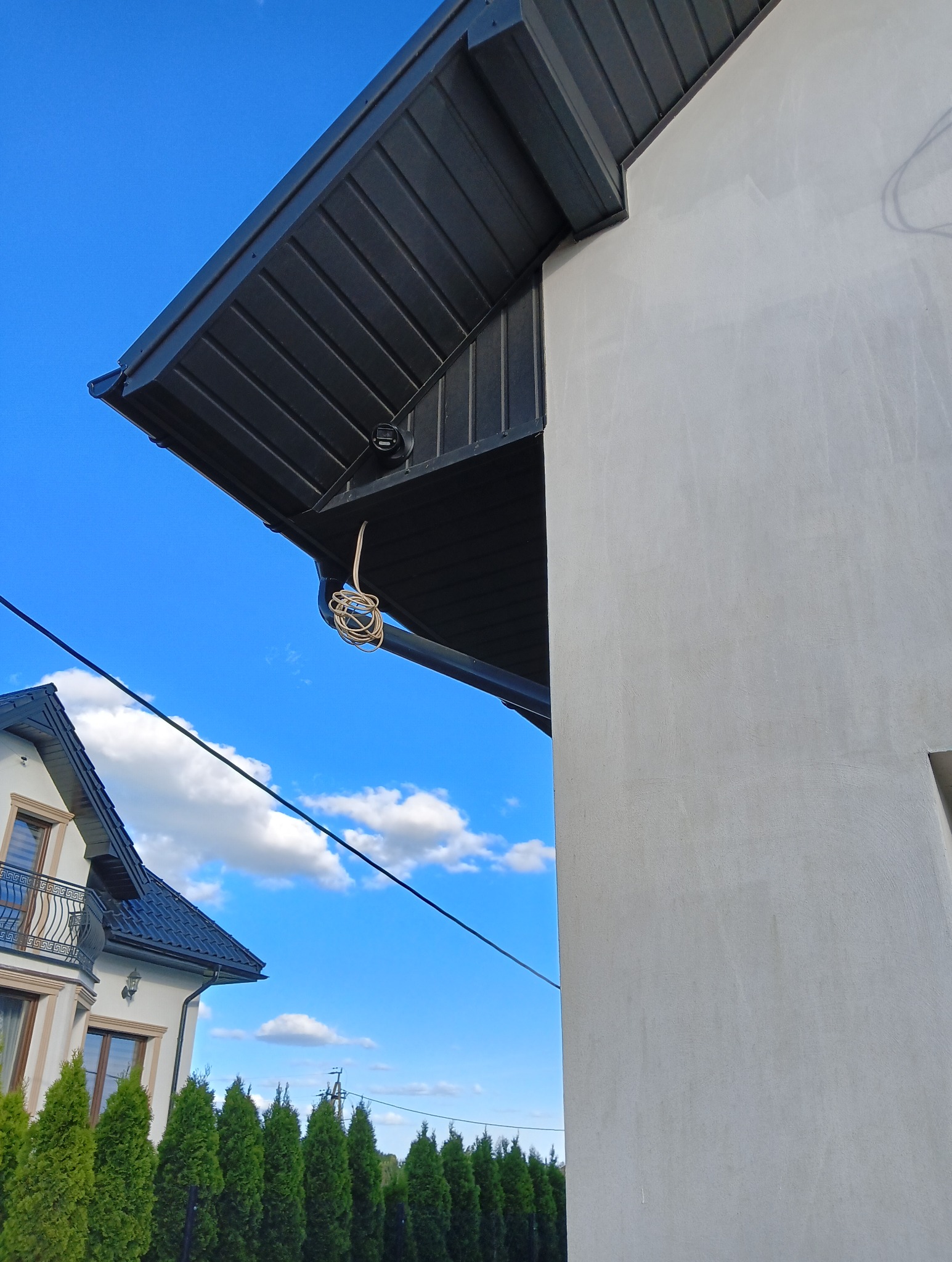 Kamera monitoringu pod czarnym okapem, z przewodem zwisającym w pętli. Fragment ściany budynku i błękitne niebo w tle. Widoczny fragment sąsiedniego domu.