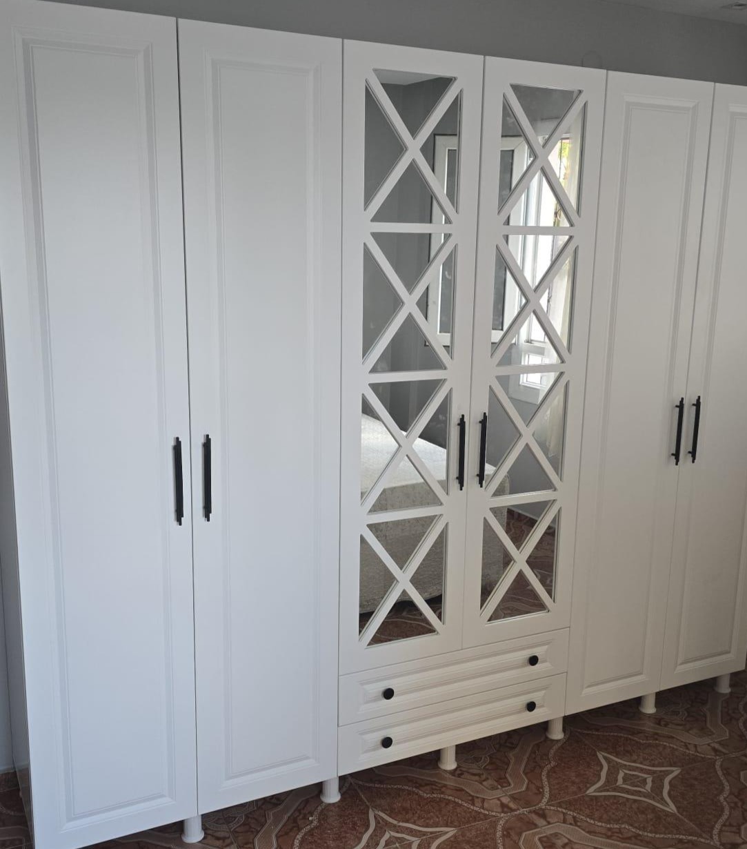 Biała szafa z lustrzanymi drzwiami i czarnymi uchwytami, widoczne dwie szuflady na dole, całość na tle brązowej podłogi. Szafa na wymiar.