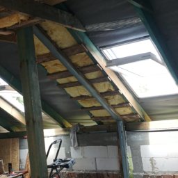 DAWID BOSIAK - Poddasze w trakcie izolacji: wełna mineralna między krokwiami, okno dachowe, widoczne elementy konstrukcji drewnianej i murowanej, drabina.