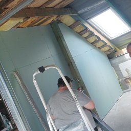 DAWID BOSIAK - Adaptacja poddasza: montaż płyt gipsowo-kartonowych przy oknie dachowym. Widoczna izolacja i metalowe profile. Praca na drabinie.