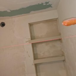 DAWID BOSIAK - Ściana po szpachlowaniu z wnękami, poziomica laserowa, wiadro z masą szpachlową na drabinie. Prace wykończeniowe w łazience.