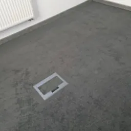 Szary dywan w biurze z widocznym włazem podłogowym i listwą przypodłogową. Grzejnik na ścianie po lewej.