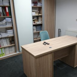 DB GROUP DAWID BOSIAK - Biuro z drewnianym biurkiem, szafą z przesuwnymi drzwiami i regałami wypełnionymi dokumentami. Na blacie leżą piloty.