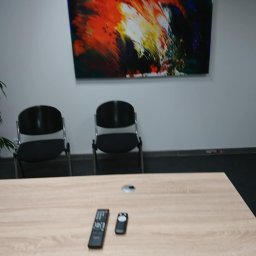 DB GROUP DAWID BOSIAK - Jasny stół konferencyjny z pilotami na tle ściany z abstrakcyjnym obrazem i dwoma krzesłami, sugerujący wnętrze biura po remoncie.