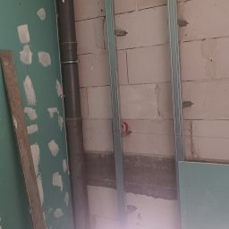 DAWID BOSIAK - Montaż stelaża pod płytę gipsową na ścianie z pustaków, widoczna rura kanalizacyjna i częściowo przytwierdzona płyta G-K. Prace wykończeniowe wewnątrz budynku.