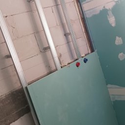 DAWID BOSIAK - Montaż płyty gipsowo-kartonowej z widoczną konstrukcją metalową i instalacją wodną (czerwony i niebieski zawór) na tle surowej ściany z pustaków.