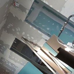 DAWID BOSIAK - Surowe ściany z widocznymi łatami szpachli, metalowe profile i płyty kartonowo-gipsowe w odcieniach beżu i zieleni, przygotowane do montażu na suficie.