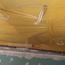 DAWID BOSIAK - Żółta folia izolacyjna pod sufitem z widocznymi elementami konstrukcyjnymi i fragmentem ściany w trakcie szpachlowania. Detal z wnętrza podczas prac wykończeniowych.