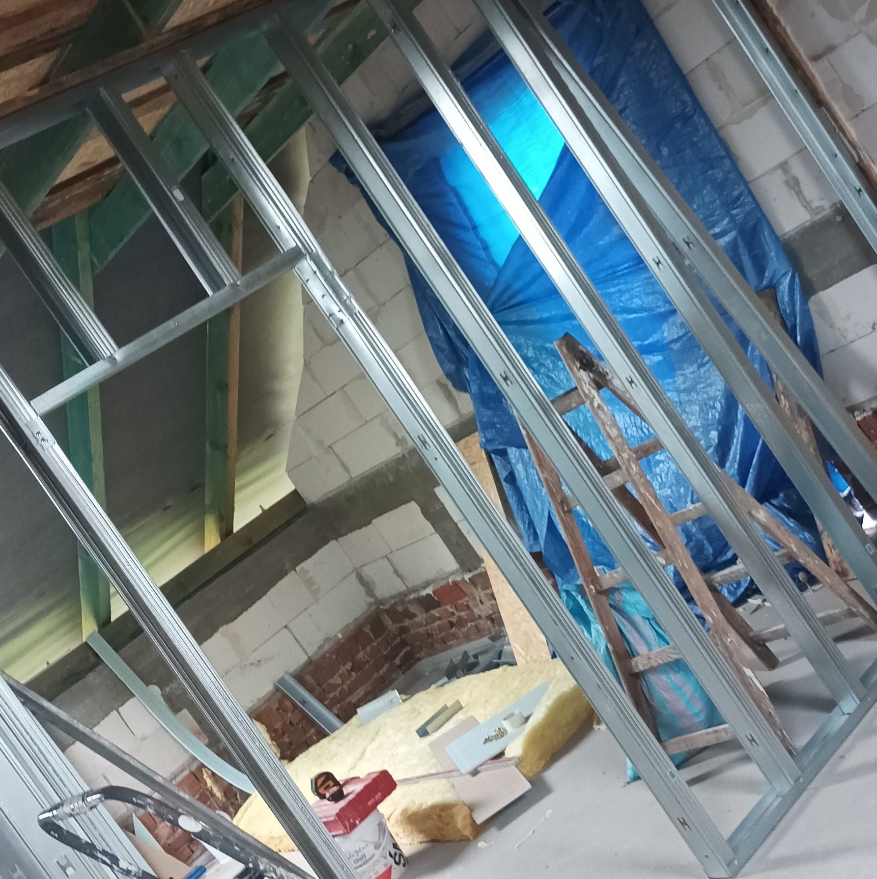 Konstrukcja metalowa pod płytę G-K na poddaszu, widoczne ocieplenie, cegła i stara drabina. Ujęcie z dołu, ukośne linie.