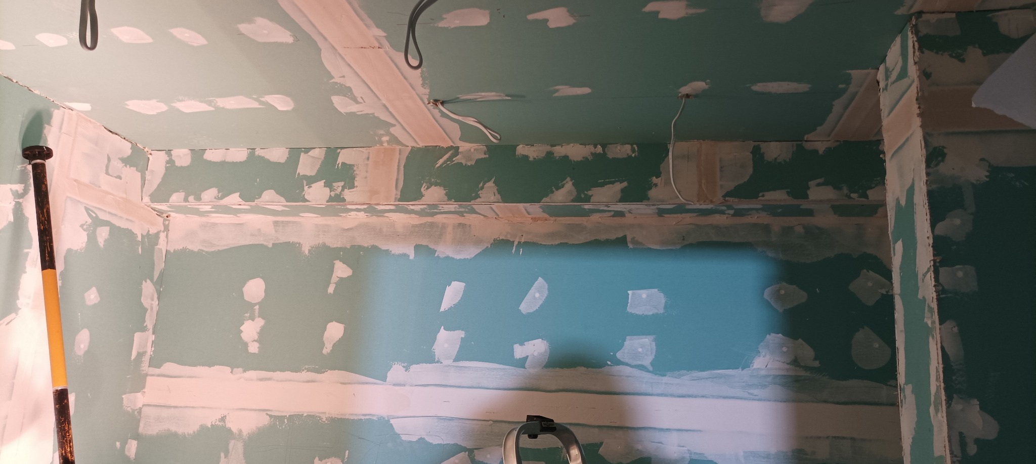 Ściana z płyt karton-gips, wstępnie szpachlowana, z widocznymi przewodami elektrycznymi i narzędziem. Wnętrze w trakcie prac wykończeniowych.