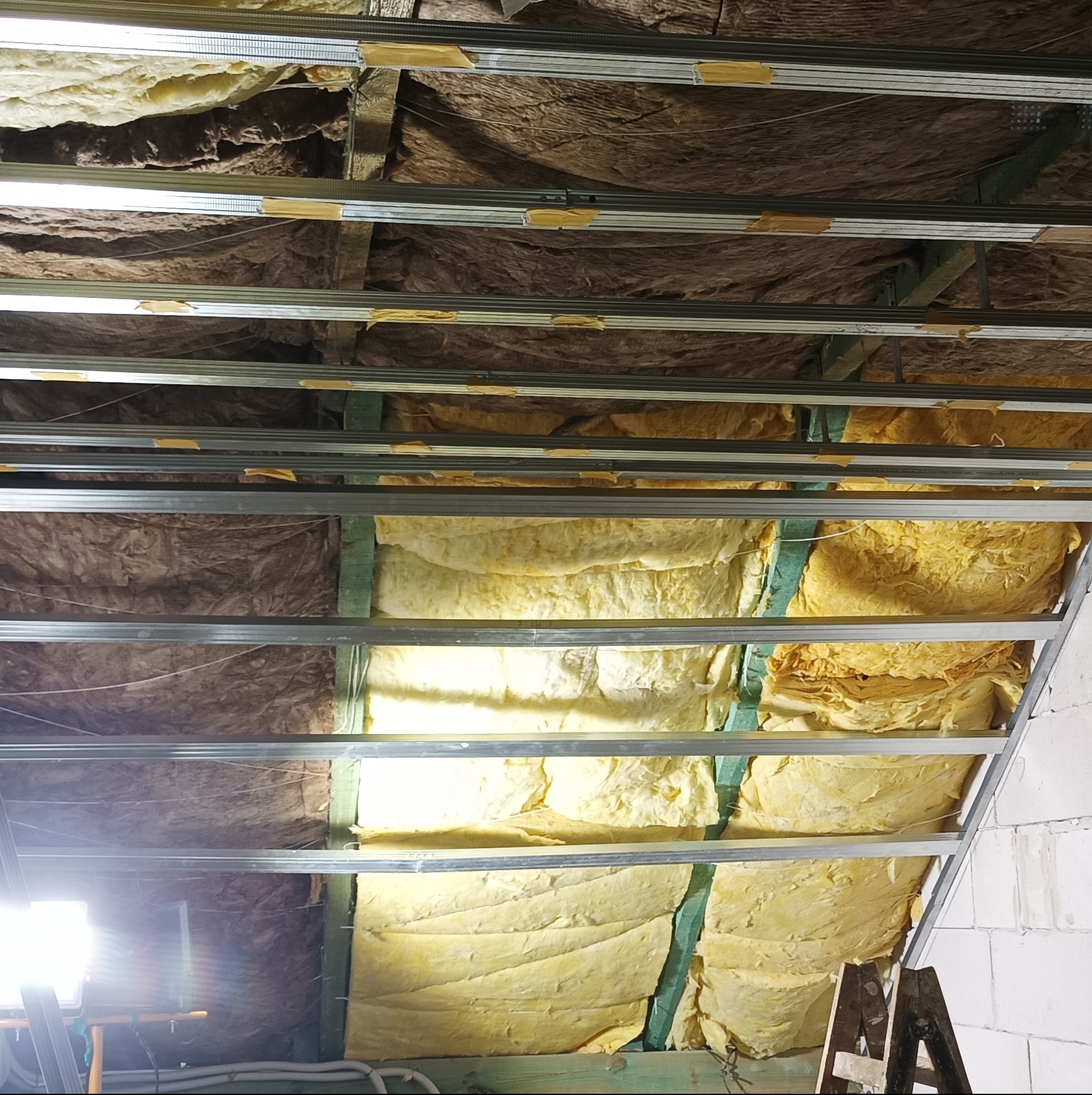 Montaż izolacji stropu wełną mineralną, widoczne profile metalowe i drewniane elementy konstrukcyjne, oświetlenie robocze, drabina. Prace wykończeniowe poddasza.