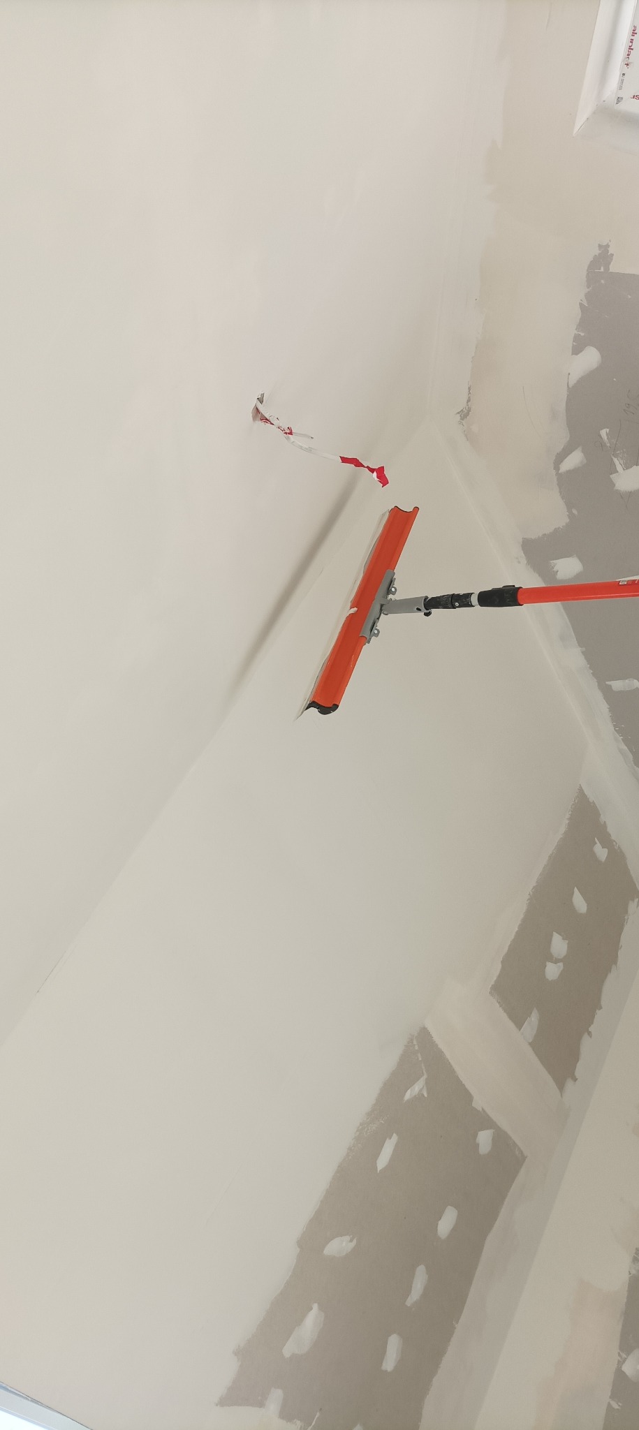 Szpachlowanie sufitu pomarańczową szpachelką na teleskopowym kiju. Widoczna zaklejona taśmą instalacja elektryczna i fragmenty niezaszpachlowanej ściany.
