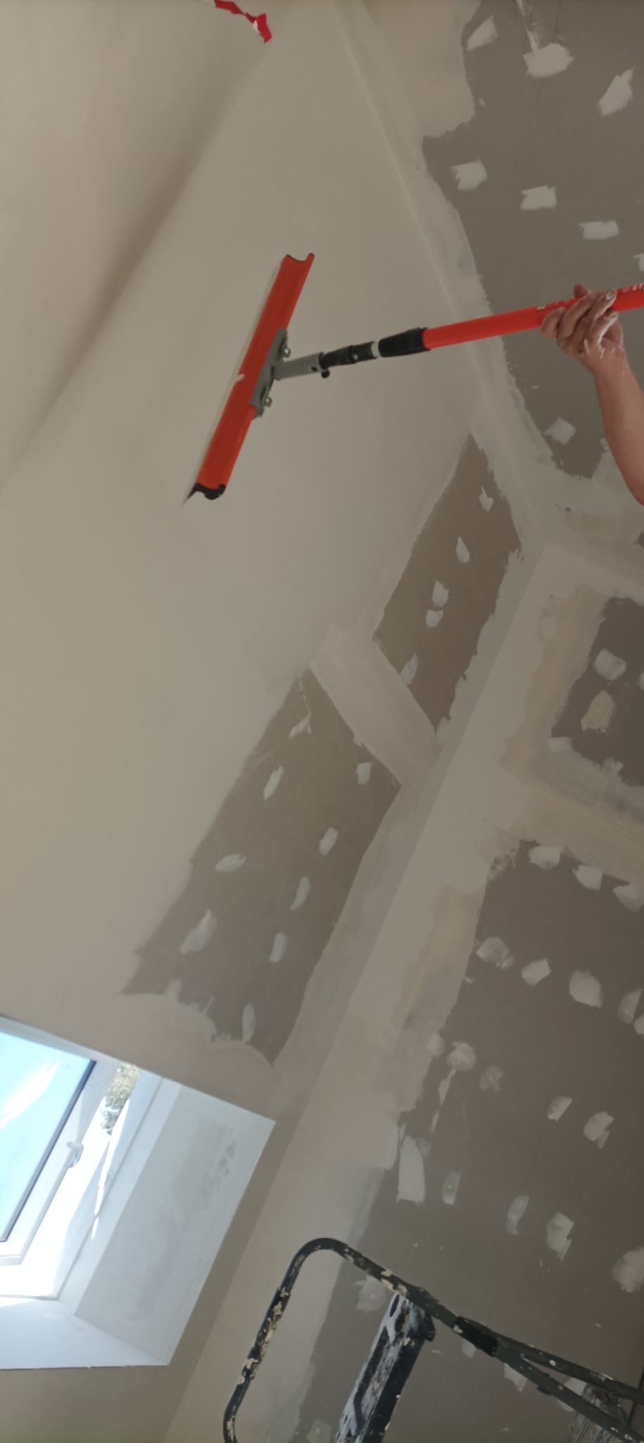 Sufit poddasza w trakcie szpachlowania, widoczne łączenia płyt gipsowych, narzędzie do szpachlowania na wysięgniku i fragment drabiny.