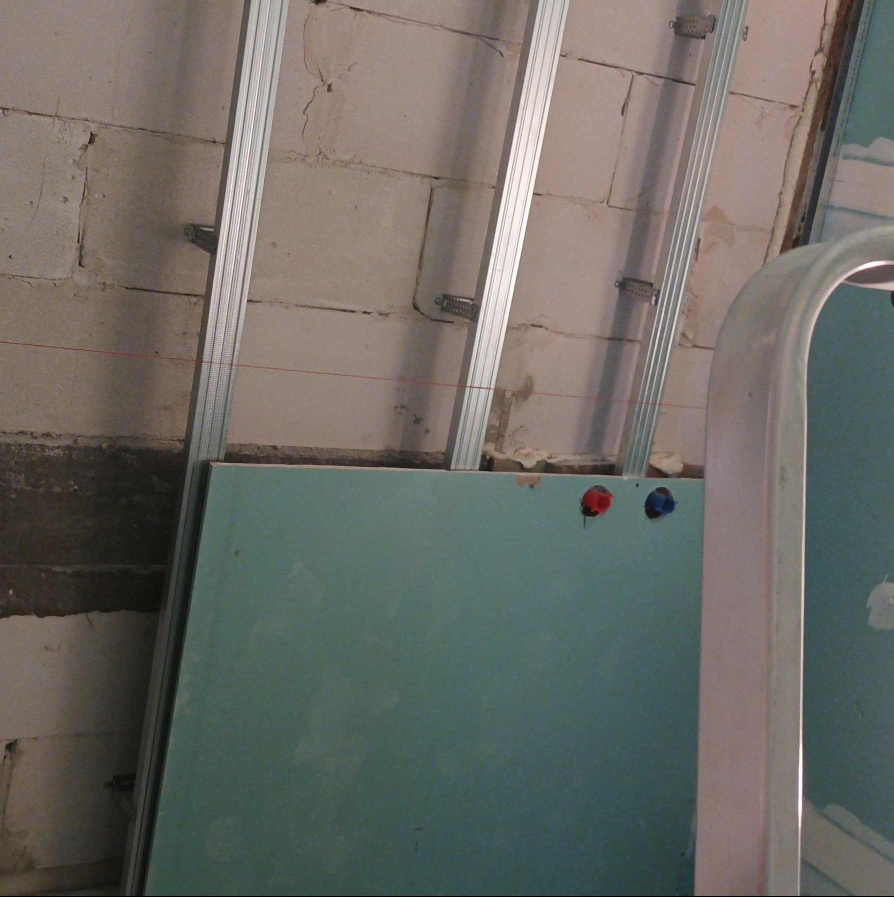 Montaż stelaża i płyt karton-gips na ścianie z pustaków, widoczne profile i instalacja elektryczna w tle. Prace wykończeniowe w trakcie realizacji.