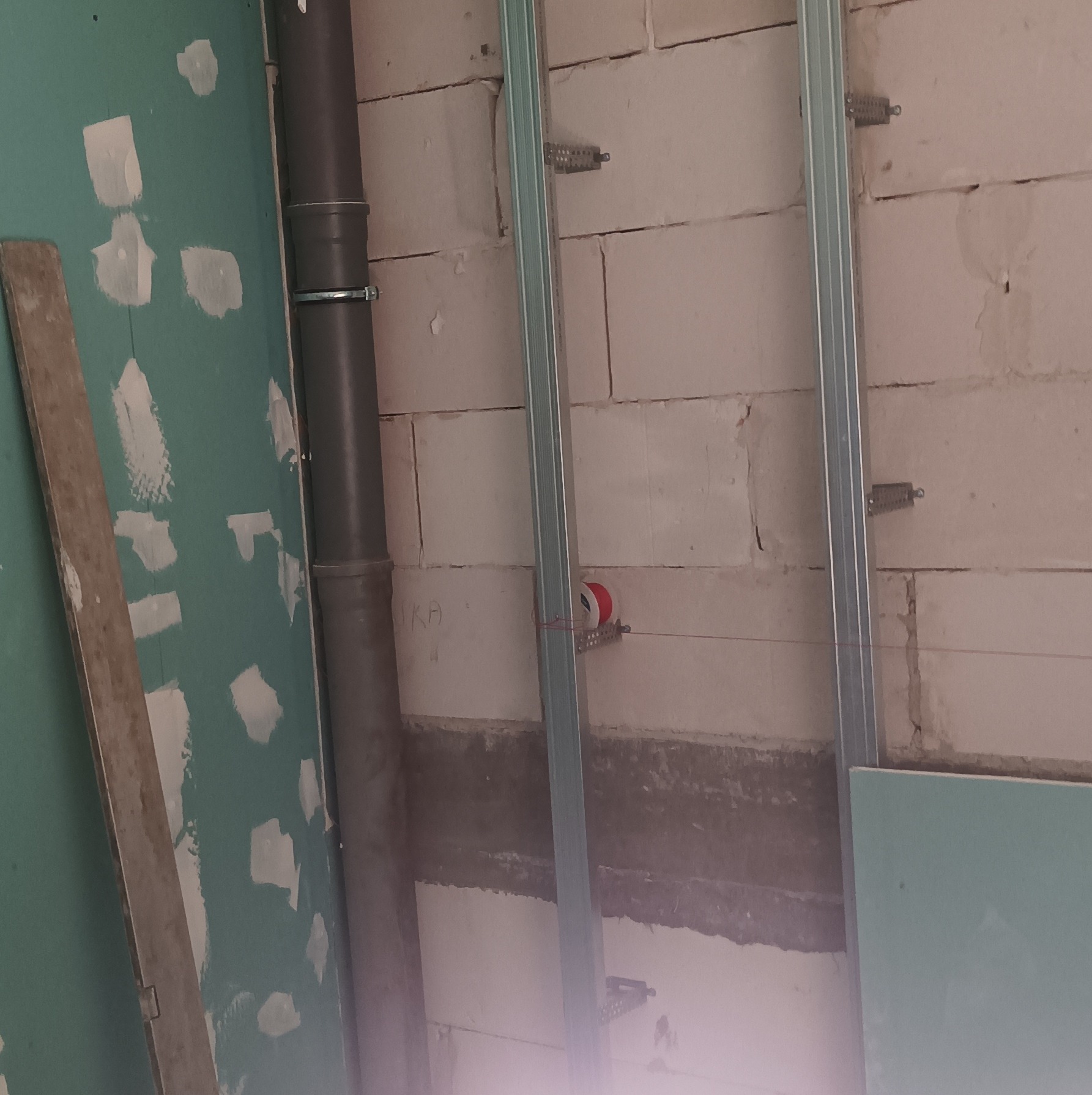 Montaż stelaża pod płytę gipsową na ścianie z pustaków, widoczna rura kanalizacyjna i częściowo przytwierdzona płyta G-K. Prace wykończeniowe wewnątrz budynku.