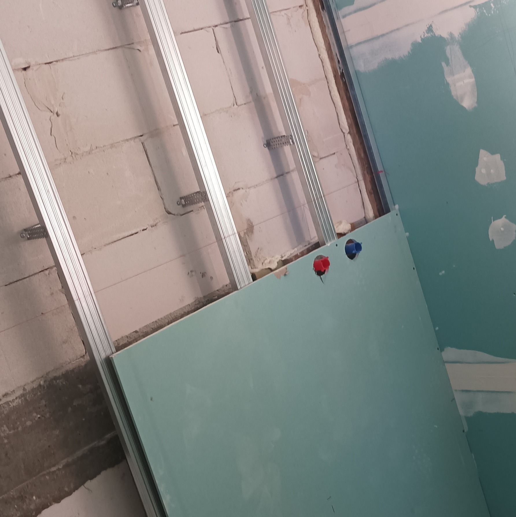 Montaż płyty gipsowo-kartonowej z widoczną konstrukcją metalową i instalacją wodną (czerwony i niebieski zawór) na tle surowej ściany z pustaków.
