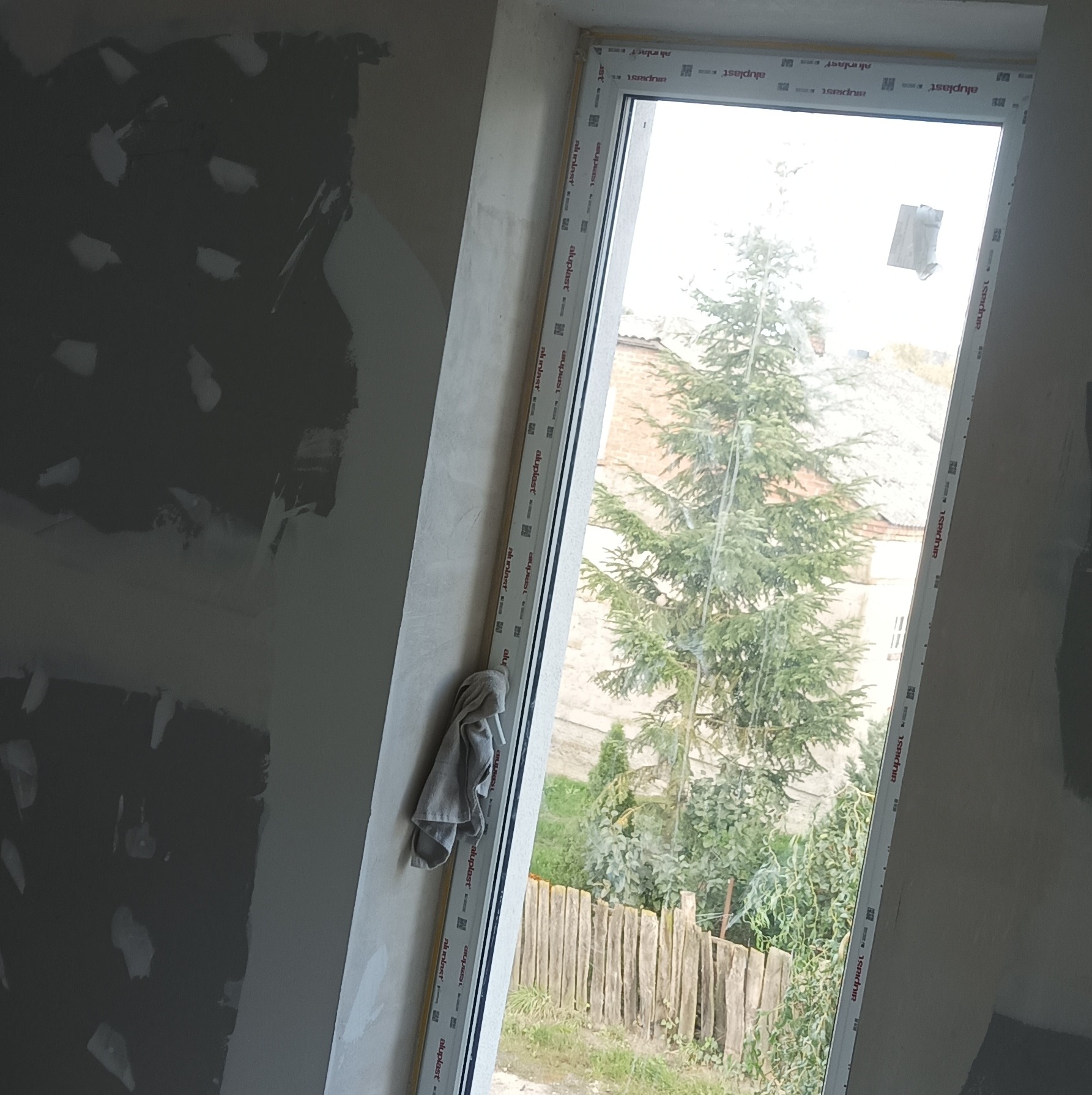 Ściana po szpachlowaniu z widocznym oknem. Na parapecie leży szmatka. Widoczne nieregularne plamy szpachli. Za oknem widok na drzewo i drewniany płot.