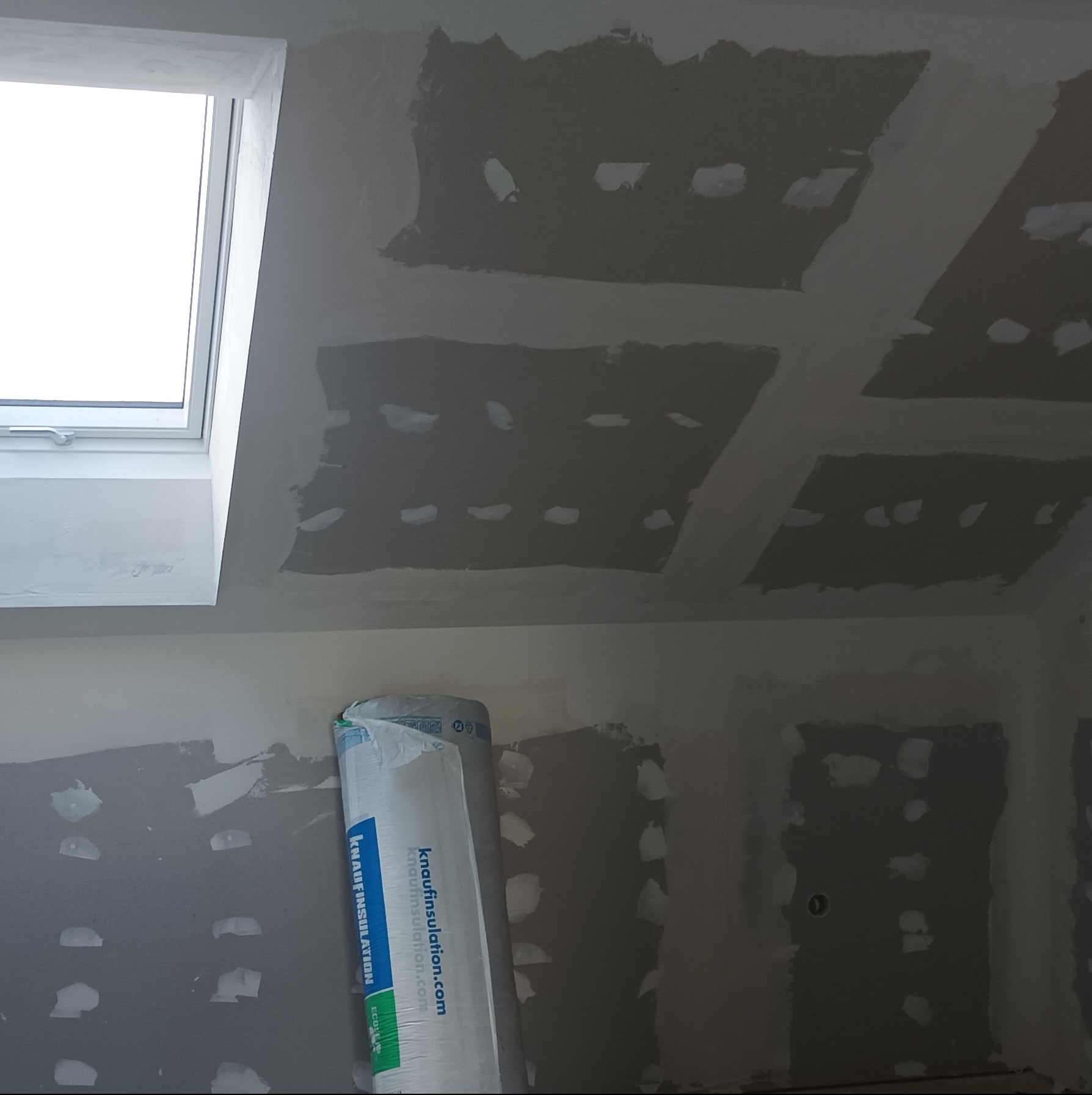 Sufit z zamontowaną płytą gipsową przed szpachlowaniem, widoczne łączenia. W rogu rolka izolacji Knauf. Okno dachowe w lewym górnym rogu.