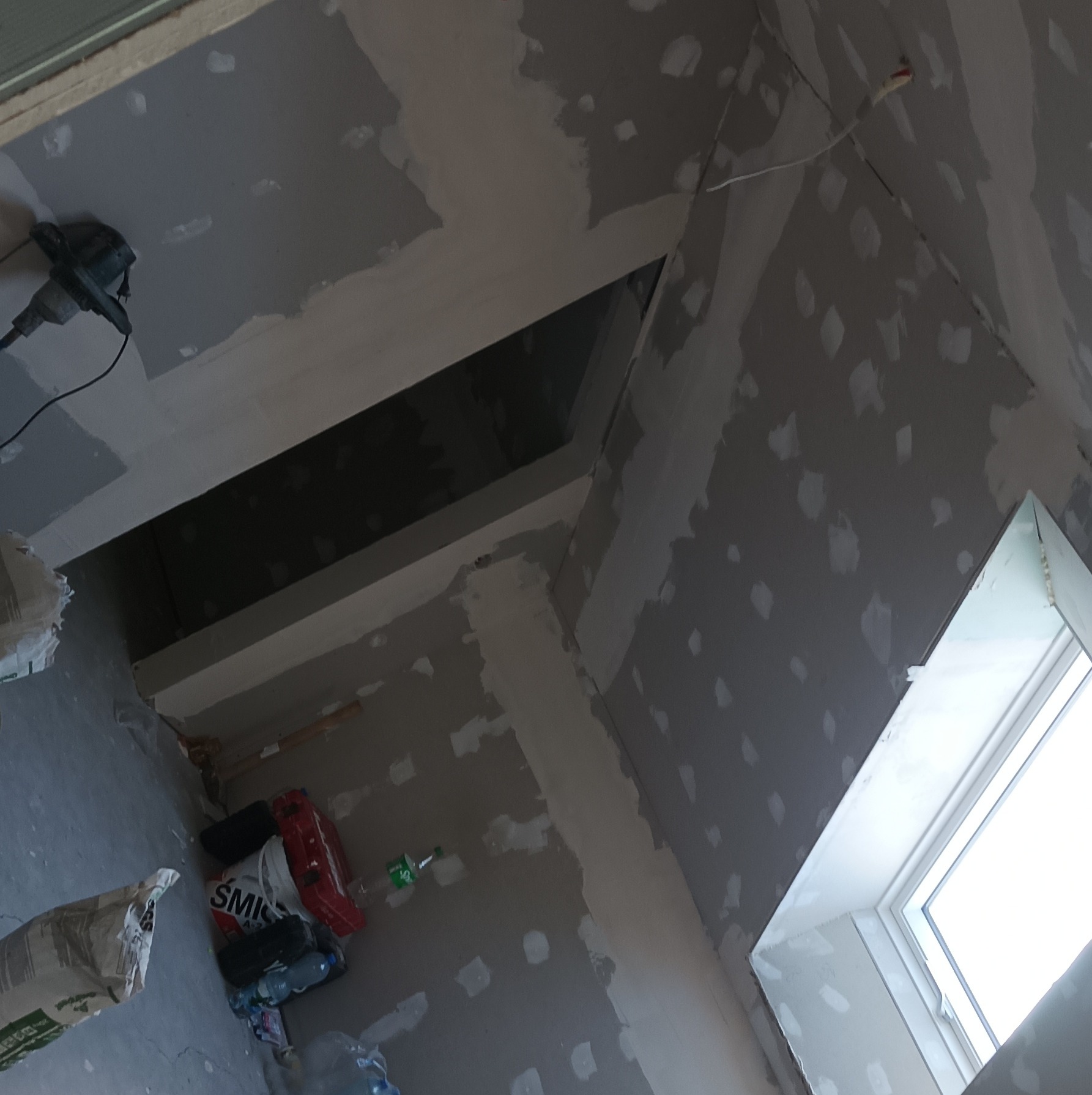 Skośne poddasze w trakcie szpachlowania, widoczne łączenia płyt gipsowych i narzędzia pracy. Ujęcie z dołu, okno dachowe w prawym rogu. W tle torba i butelki.