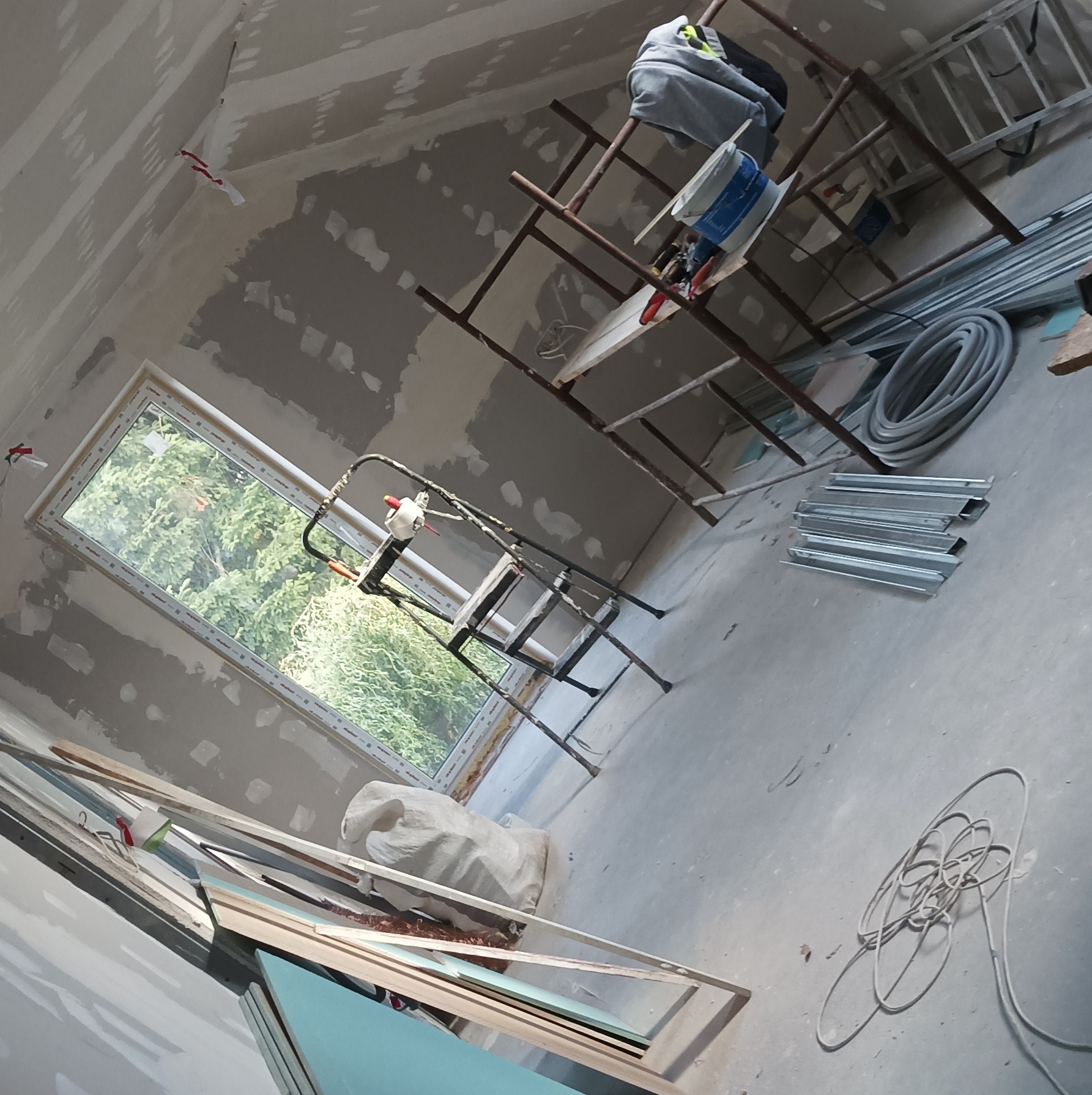Skośne poddasze w trakcie szpachlowania, widoczne rusztowania, okno z widokiem na zieleń i materiały budowlane. Ściany częściowo pokryte szpachlą.