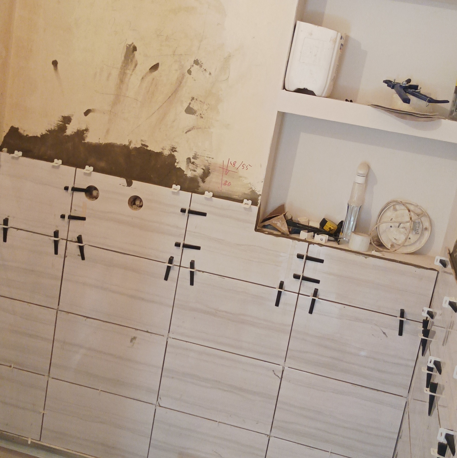 Układanie jasnych, marmurkowych płytek na ścianie z użyciem klipsów poziomujących. Widoczne narzędzia i fragment ściany z resztkami zaprawy.