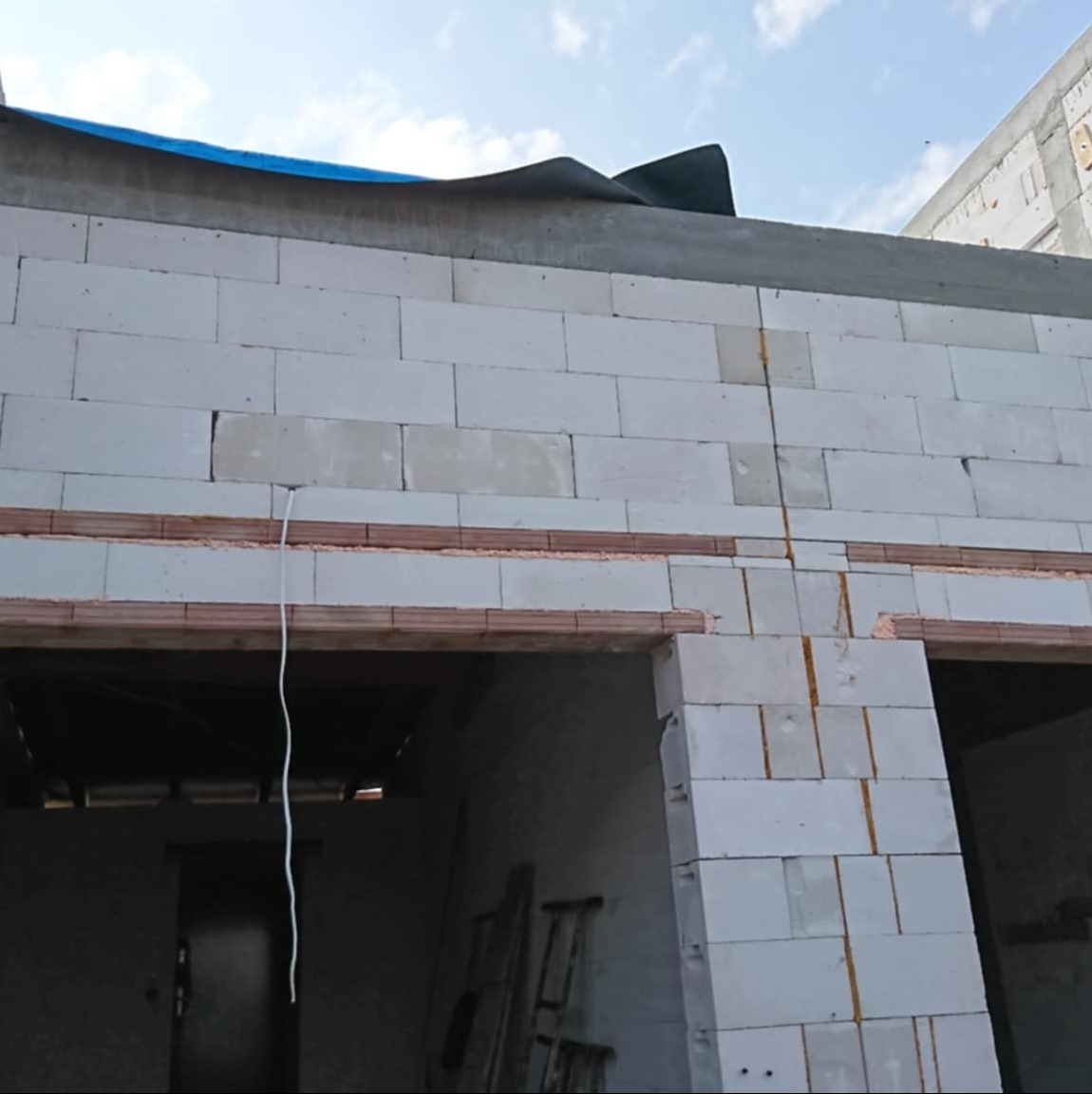 Ściana z białych bloczków, widoczne otwory okienne z drewnianymi nadprożami, sznur pionujący. Budowa domu w trakcie realizacji.