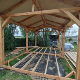 Konstrukcja drewnianej altany w trakcie budowy; widoczne elementy szkieletu dachu i podłogi na czarnej macie. Tło: zieleń ogrodu, częściowo zadaszone miejsce.