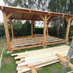 Tomasz Hajzler inwestycje budowlane - Drewniana konstrukcja z zadaszeniem w trakcie budowy na zewnątrz. Widoczne deski, drabina i narzędzia. Tło stanowią drzewa i zieleń. Ujęcie z dołu.