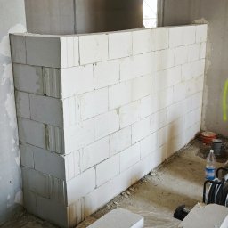 Tomasz Hajzler inwestycje budowlane - Wnętrze w trakcie budowy z częściowo wymurowaną ścianką działową z białych bloczków, poziomica leży na bloczkach. Widoczne surowe ściany i podłoga.