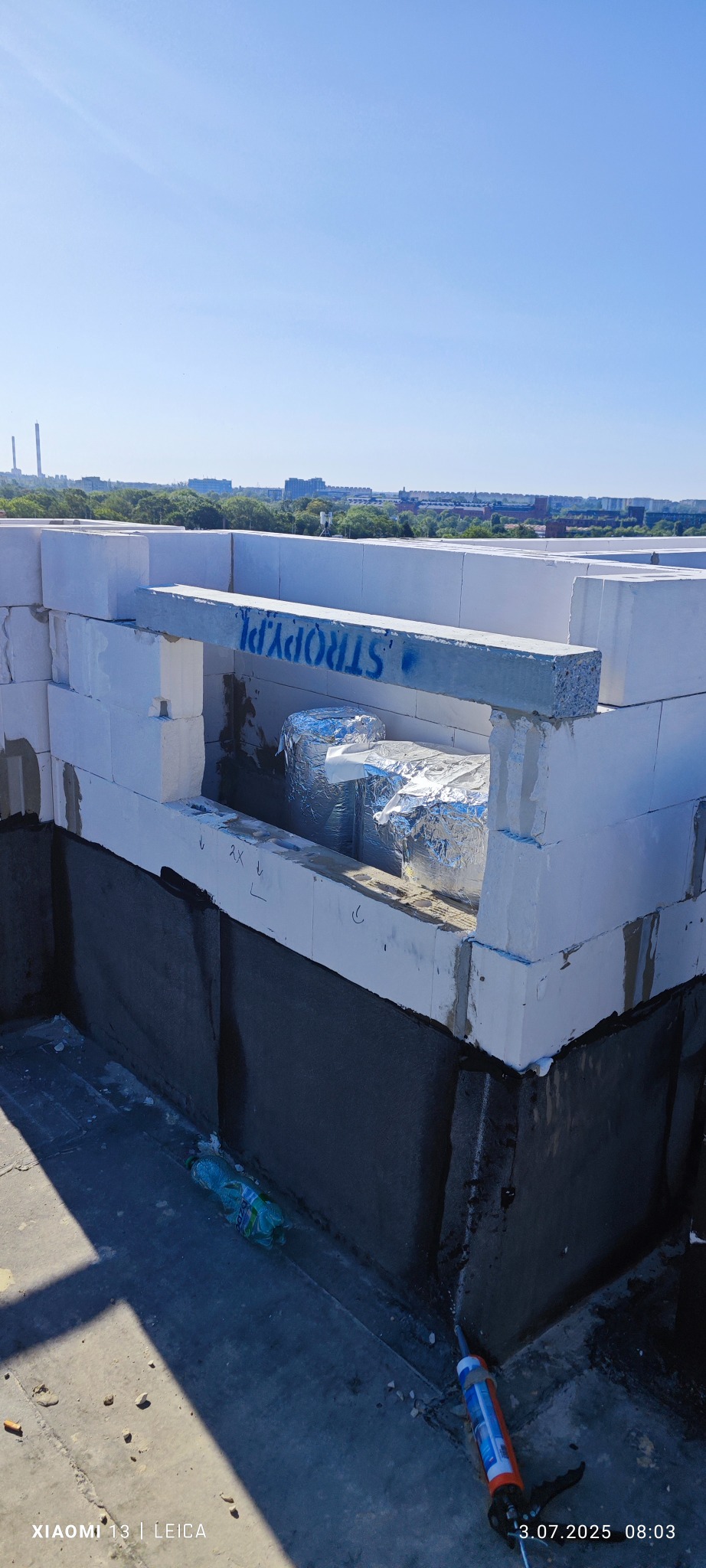 Widok na mur z betonu komórkowego z otworem na nadproże, zabezpieczony hydroizolacją. W tle zabudowa miejska i błękitne niebo. Na pierwszym planie butelka i aplikator do uszczelniacza.